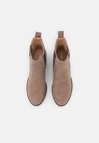 Une paire de bottes en suède taupe à hauteur de cheville avec des panneaux élastiques noirs sur les côtés et des tirettes, présentée en vue de dessus sur un fond blanc.