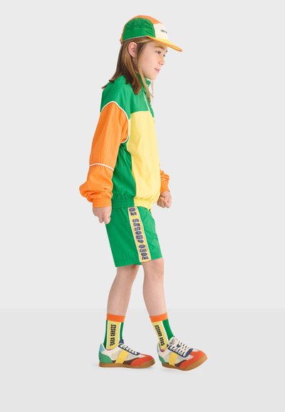 Enfant debout de profil portant une veste multicolore verte, jaune et orange, un short assorti, une casquette, des chaussettes orange et des baskets multicolores.