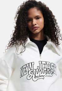 Felpa bianca con zip e dettagli neri "NEW JERSEY Athletic". Presenta un colletto alto e una texture morbida, adatta per un abbigliamento informale.