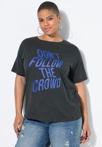 Persona segura de sí misma con cabello corto, usando pendientes de aro, una camiseta negra con el texto "NO SIGAS A LA MULTITUD" y jeans azules rasgados.