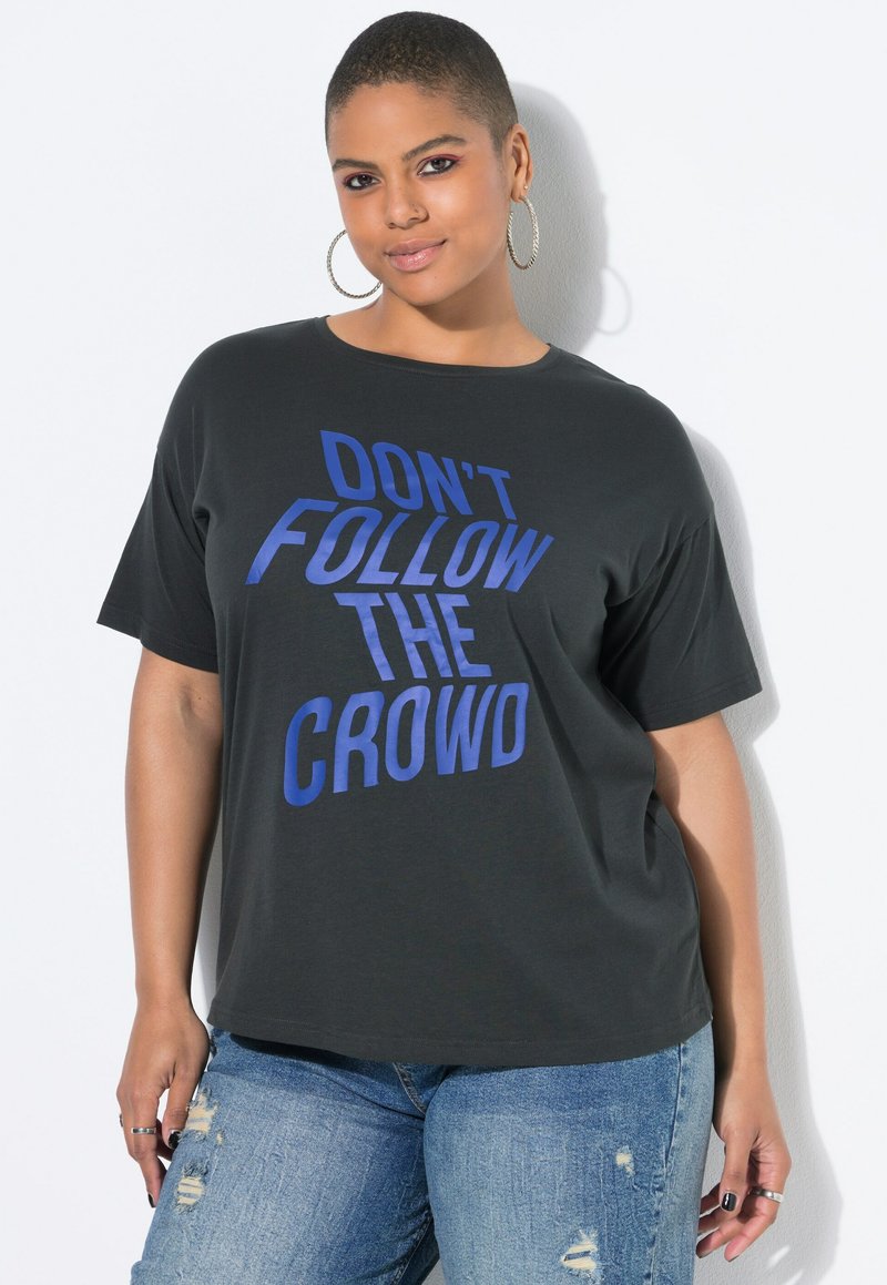 Persona segura de sí misma con cabello corto, usando pendientes de aro, una camiseta negra con el texto "NO SIGAS A LA MULTITUD" y jeans azules rasgados.