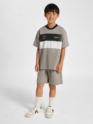 Junge steht, trägt taupefarbene Shorts und ein passendes kurzärmeliges Hemd mit schwarz-weißen horizontalen Streifen, weiße Socken und beige Turnschuhe.