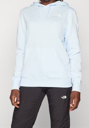 Persoon die een lichtblauwe The North Face-hoodie en zwarte The North Face-broek draagt, staand met ontspannen handen langs het lichaam.