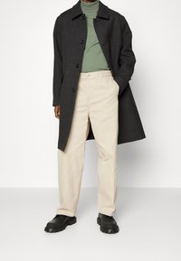 Persoon in een donkergrijze lange jas, groene coltrui, beige broek en zwarte chunky schoenen, staand tegen een witte achtergrond.