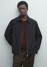 Black zip-up jacket with a pointed collar, worn over a rust-colored shirt, featuring a smooth texture and minimalistic design.