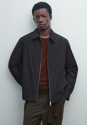 Giacca nera con zip e colletto a punta, indossata sopra una camicia color ruggine, caratterizzata da una texture liscia e un design minimalista.