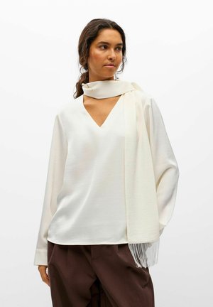 Une femme porte un chemisier blanc à encolure en V, assorti d’une écharpe à franges et d’un pantalon marron foncé, sur un fond de studio uni.