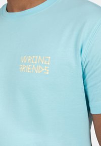 Camiseta de algodón azul claro con mangas cortas y cuello acanalado. Presenta la inscripción "WRONG FRIENDS" en letras doradas en relieve en la parte delantera.