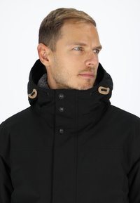 Black jacket with a high collar, button front, and textured inner lining. Features contrasting tan accents on collar and buttons.