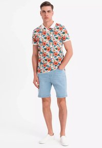 Polo shirt floral avec des motifs orange et verts, col blanc, associé à un short en denim clair et des chaussures blanches à enfiler.