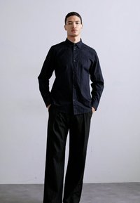 Svart teksturert skjorte med knapplukking og lange ermer, paret med løse svarte pinstripete bukser, vist på en modell mot en lys bakgrunn.