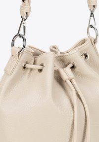 Sac seau en cuir crème avec surface texturée, fermeture à cordon, et accents en métal argenté. Dispose de sangles lisses et d'œillets ronds.