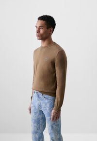 Suéter marrón de punto con cuello redondo, con un pequeño logo verde. Combinado con jeans de patrón azul claro. Textura suave.