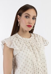 Blusa beige con maniche a cappuccio arricciate, ricamo floreale argentato, dettagli plissettati sul davanti, abbinata a orecchini a cerchio in perle.