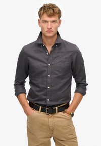 Camisa de botonadura oscura de color gris con un cuello, textura sutil y un bolsillo en el pecho, combinada con pantalones caqui y un cinturón negro.