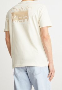 Camiseta de algodón color crema con un gran logo "Calvin Klein" desgastado en la parte de atrás. Mangas cortas y un clásico escote redondo.