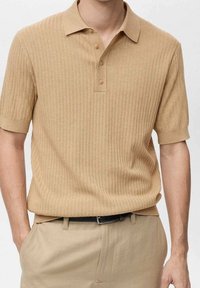 Polo beige à côtes avec des manches courtes, col à deux boutons et tissu texturé, associé à un pantalon beige assorti et une ceinture noire.