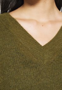 Gros plan sur une personne portant un pull en maille vert olive à col en V, montrant le cou et une partie des cheveux foncés.