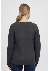 Mujer con cabello rubio en una coleta, vistiendo un suéter de manga larga texturizado gris oscuro y jeans azules, vista desde atrás sobre un fondo blanco.