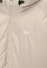 Veste beige avec fermeture éclair frontale, capuche à cordon de serrage et petit logo brodé "Swtr" sur la poitrine.