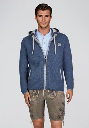 Mann trägt einen blauen Kapuzenpullover mit Reißverschluss, ein hellblaues Hemd und traditionelle Lederhosen mit Stickerei, steht vor weißem Hintergrund.