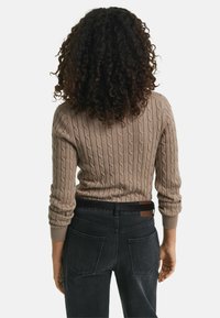 Beige Kabelstrickpullover mit tailliertem Design und gerippten Bündchen, kombiniert mit dunklen Jeans mit zwei Gesäßtaschen und einem Gürtelakzent.