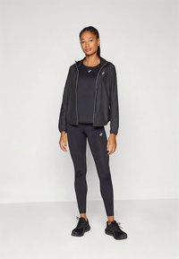 Veste de sport noire avec fermeture éclair et accents blancs, associée à un haut noir moulant et des leggings, tous deux en matériau extensible. Baskets noires.