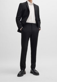 Czarny garnitur o dopasowanym kroju, z jednorzędową marynarką, białą koszulą pod spodem i czarnymi skórzanymi butami. Gładka faktura.