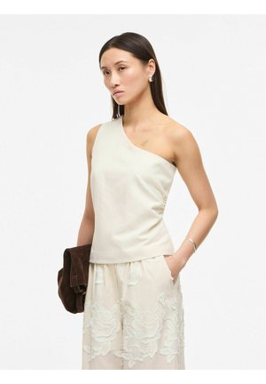 Kobieta w białej bluzce odsłaniającej ramiona i jasnej spódnicy zdobionej kwiatowym haftem, trzyma ciemnobrązową zamszową torbę, jedną ręką trzymając w kieszeni.