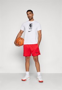 Men's sports outfit featuring a white t-shirt with NBA logo, red mesh shorts with "BULLS" text, and white sneakers with red accents, holding a basketball.