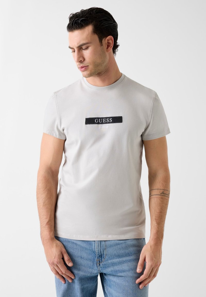 T-shirt en coton gris clair avec un patch logo rectangulaire noir "GUESS" sur la poitrine ; manches courtes et coupe ajustée.