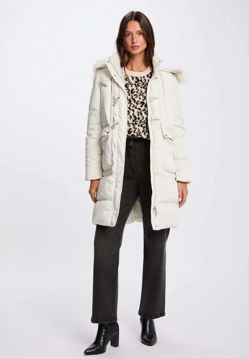 Cappotto imbottito bianco con cappuccio e bordo in pelliccia, chiusure a bottone e grandi tasche, indossato sopra un maglione con stampa leopardata e jeans neri.