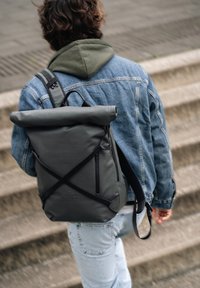 Zaino grigio a sacca con una texture liscia, spallacci regolabili, tasca frontale con zip e dettagli del logo, indossato su una giacca di jeans.