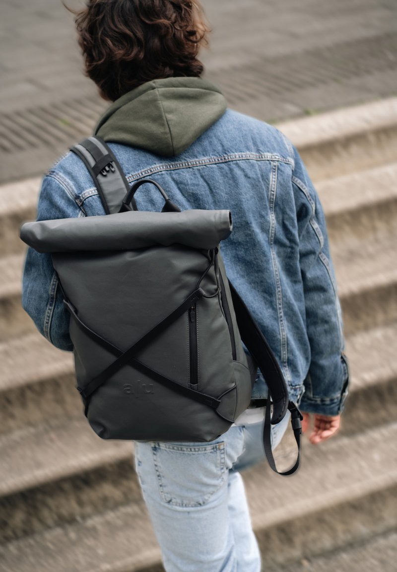 Zaino grigio a sacca con una texture liscia, spallacci regolabili, tasca frontale con zip e dettagli del logo, indossato su una giacca di jeans.