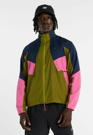 Uomo che indossa un giubbotto a vento color-block con pannelli oliva, navy e rosa, pantaloncini neri e un cappellino nero New Balance, in piedi davanti a uno sfondo semplice.