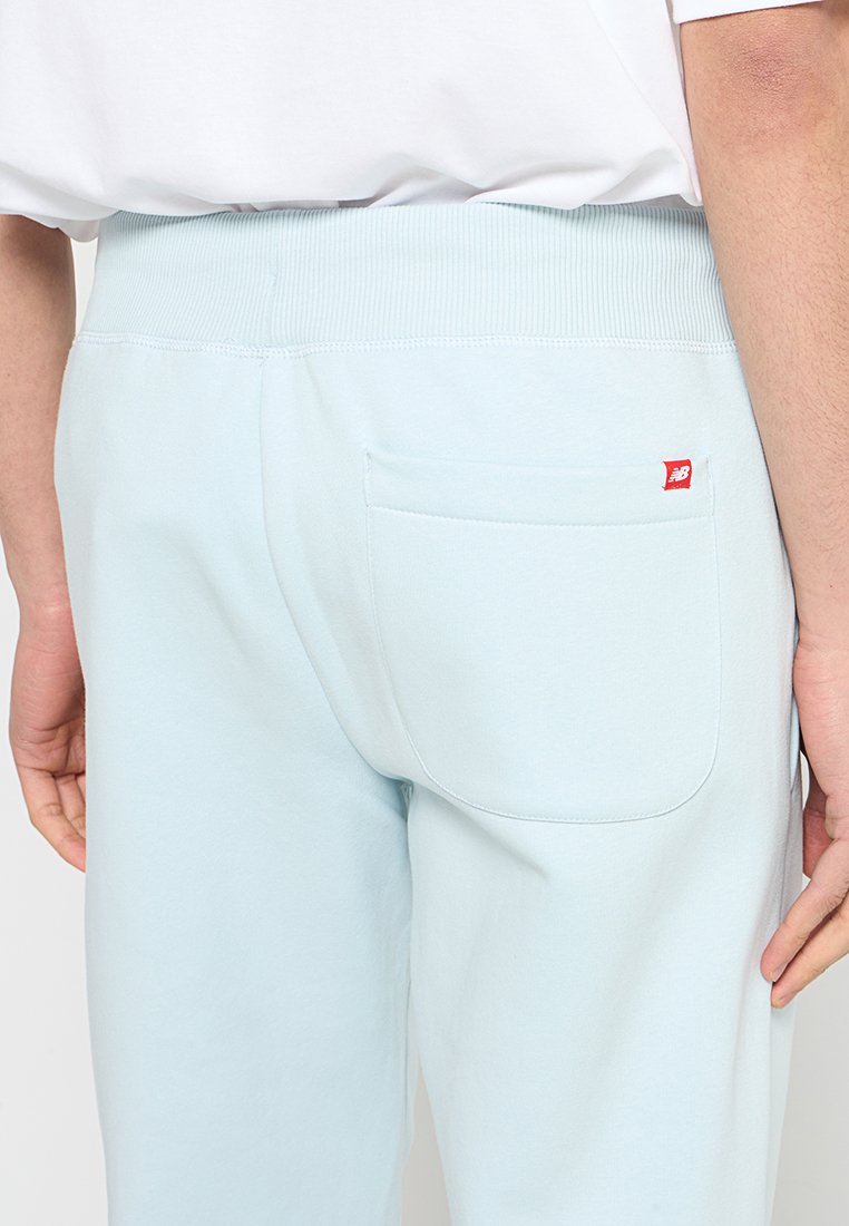 Pantalon de sport bleu clair avec une taille élastique à côtes et une poche plaquée à l'arrière, arborant une petite étiquette de logo rouge. Texture lisse.
