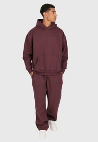 Junger Mann steht mit den Händen in den Taschen, trägt einen passenden dunkelburgunderfarbigen Kapuzenpullover und Jogginghosen, schaut nach rechts, weiße Sneakers.