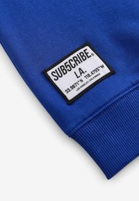 Kungligt blå sweatshirttyg med ribbade muddar. Har en vit rektangulär etikett med svart text som anger varumärke och koordinater.
