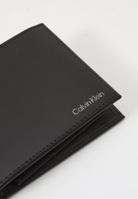 Svart läderplånbok med en slät yta, med sydda kanter och en silverfärgad "Calvin Klein"-logotyp i det nedre högra hörnet.