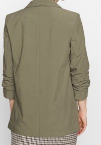 Blazer vert olive avec un design structuré, manches longues et poignets retroussés, présentant une texture lisse et des lignes épurées.