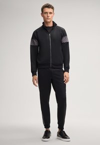 Zwarte zip-up trainingsjack met gestructureerde grijze accenten op de schouders, gecombineerd met bijpassende zwarte tapered broek en zwarte sneakers.