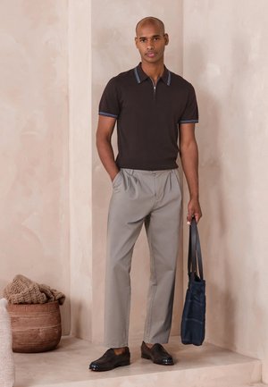 Muž oblečený v čiernom tričku s krátkym rukávom a svetlosivých nohaviciach, držiaci námornícku tašku, stojí v béžovej miestnosti vedľa pleteného koša s dekou.