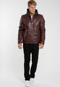 Burgundy läderjacka med frontdragkedja, två dragkedjefickor på bröstet och en grå hoodie under, ihop med svarta byxor och vita sneakers.