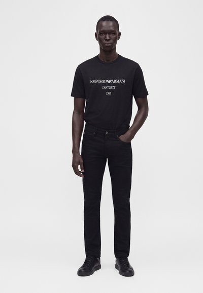 Άνδρας που φοράει μαύρο μπλουζάκι Emporio Armani, μαύρα τζιν και μαύρα αθλητικά παπούτσια, στέκεται με το ένα χέρι στην τσέπη σε απλό λευκό φόντο.