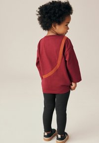 Chemise bordeaux à manches longues avec un accent orange le long de l'épaule, associée à des leggings noirs côtelés et des chaussures foncées avec des semelles beiges.