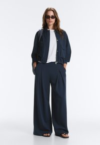 Femme portant des lunettes de soleil noires, une veste bleu marine avec de grandes poches, une chemise blanche, un pantalon large bleu marine et des sandales noires, debout les mains dans les poches.