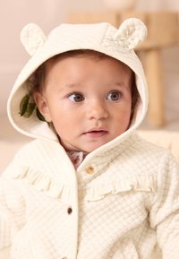 Veste à capuche matelassée couleur crème avec oreilles d'ours, accents froncés et fermeture à boutons. Texture douce avec une doublure rose clair visible.
