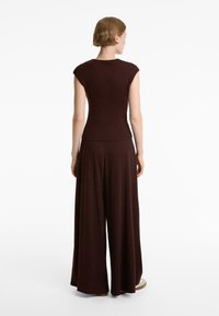 Top marrone aderente senza maniche con una texture liscia, abbinato a pantaloni a gamba larga. L'outfit presenta un design minimalista e un colore solido.