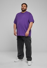 Lilla oversized t-skjorte med kortermede ermer, kombinert med mørkegrå cargobukser med lommer, og hvite joggesko. Enkelt bakgrunn.
