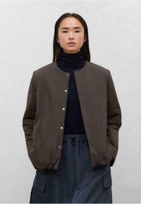 Chaqueta marrón de botón con una textura suave, cuello redondo y cierres a presión, llevada sobre un cuello alto oscuro y combinada con una falda azul.